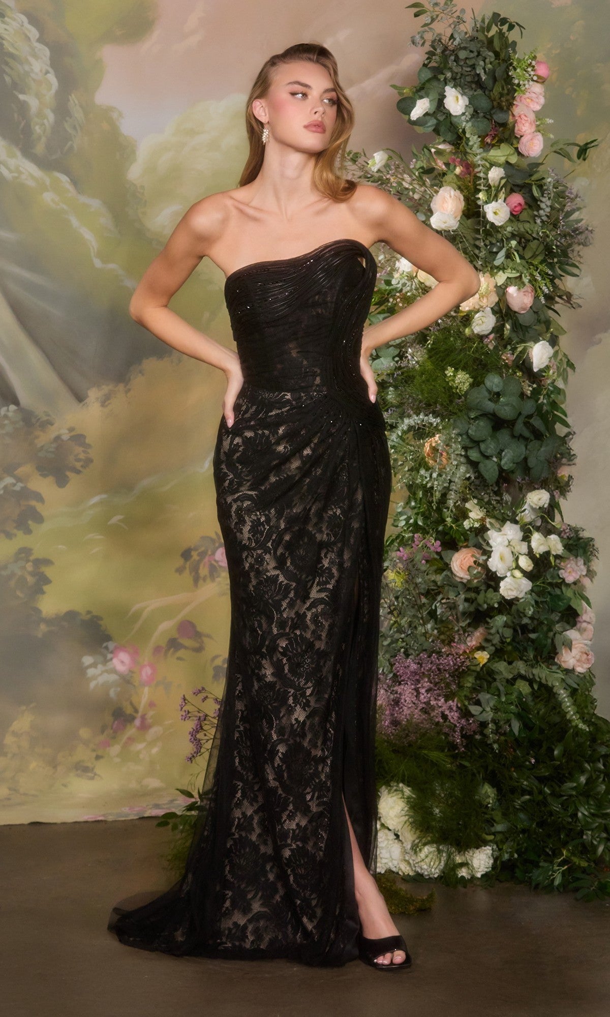 A woman wearing the black Andrea and Leo A1497 long prom dress stands with hands on hips in front of a floral arrangement and painted backdrop.